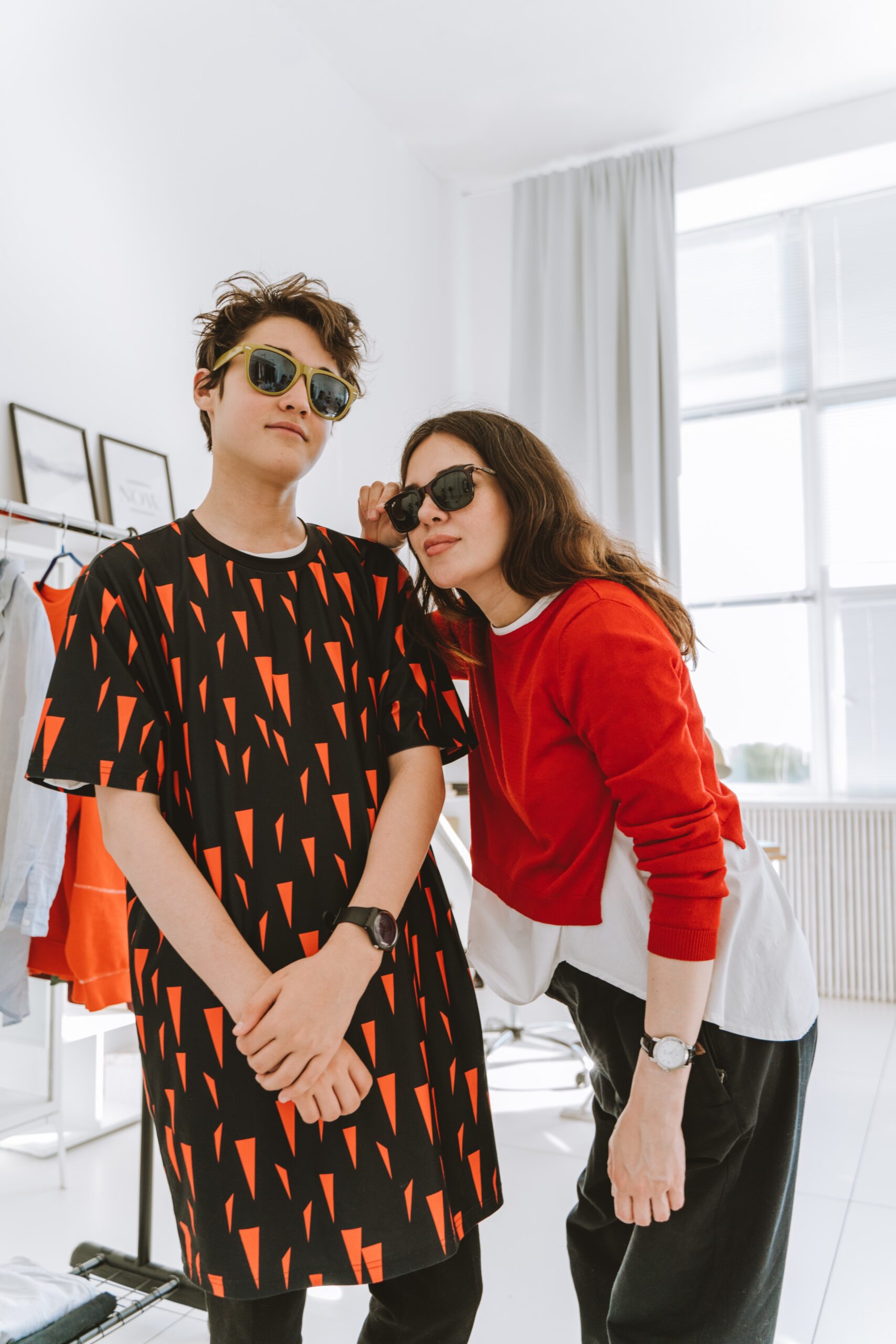 Two cool teens posing in their show costumes in a changing room that has a bright window with white curtains that are flung open and both are wearing sunglasses.