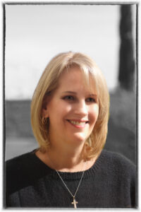 Suzanne Allee's Head Shot photo in black and white wearing a blouse with her necklace of a cross showing