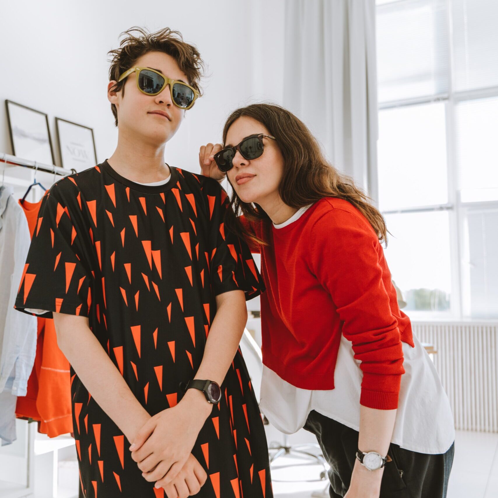Two cool teens posing in their show costumes in a changing room that has a bright window with white curtains that are flung open and both are wearing sunglasses.