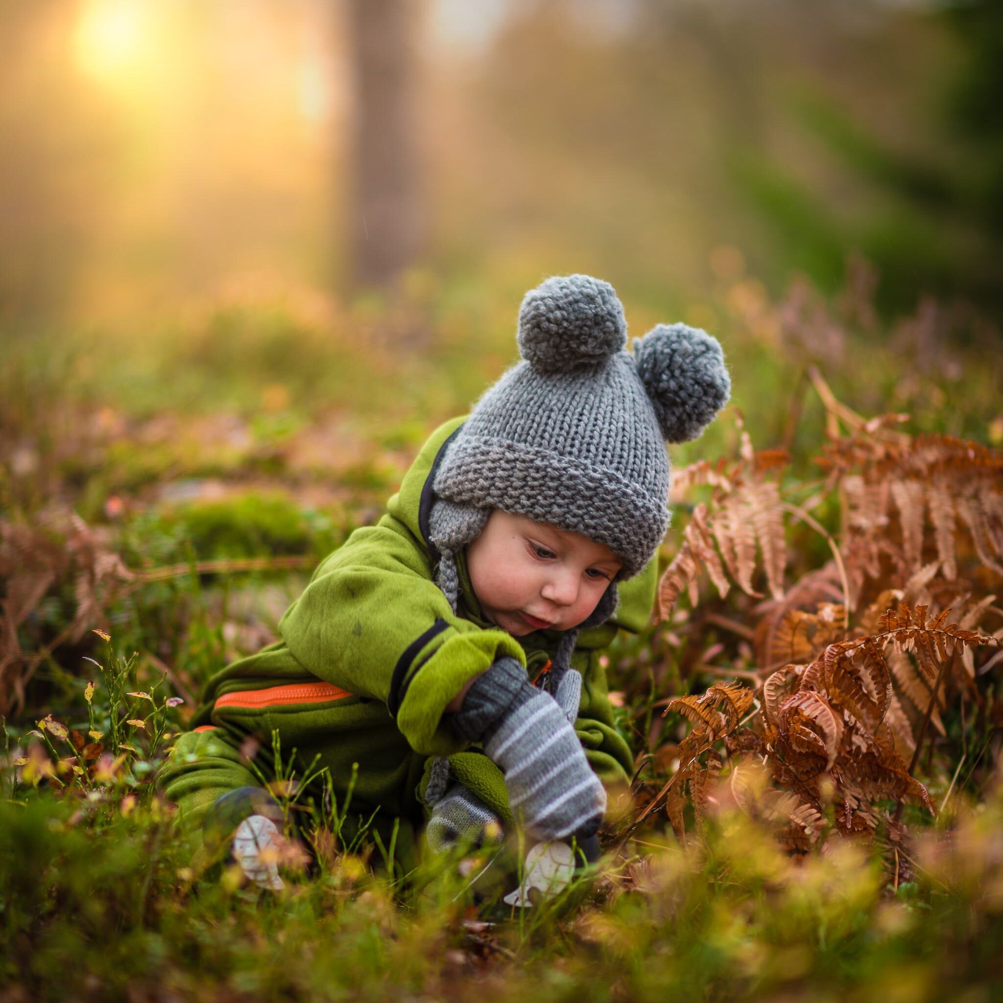 A young baby sitting in the grass wearing gloves and a knitted cap. It's during the early fall with no weather but color are changing.