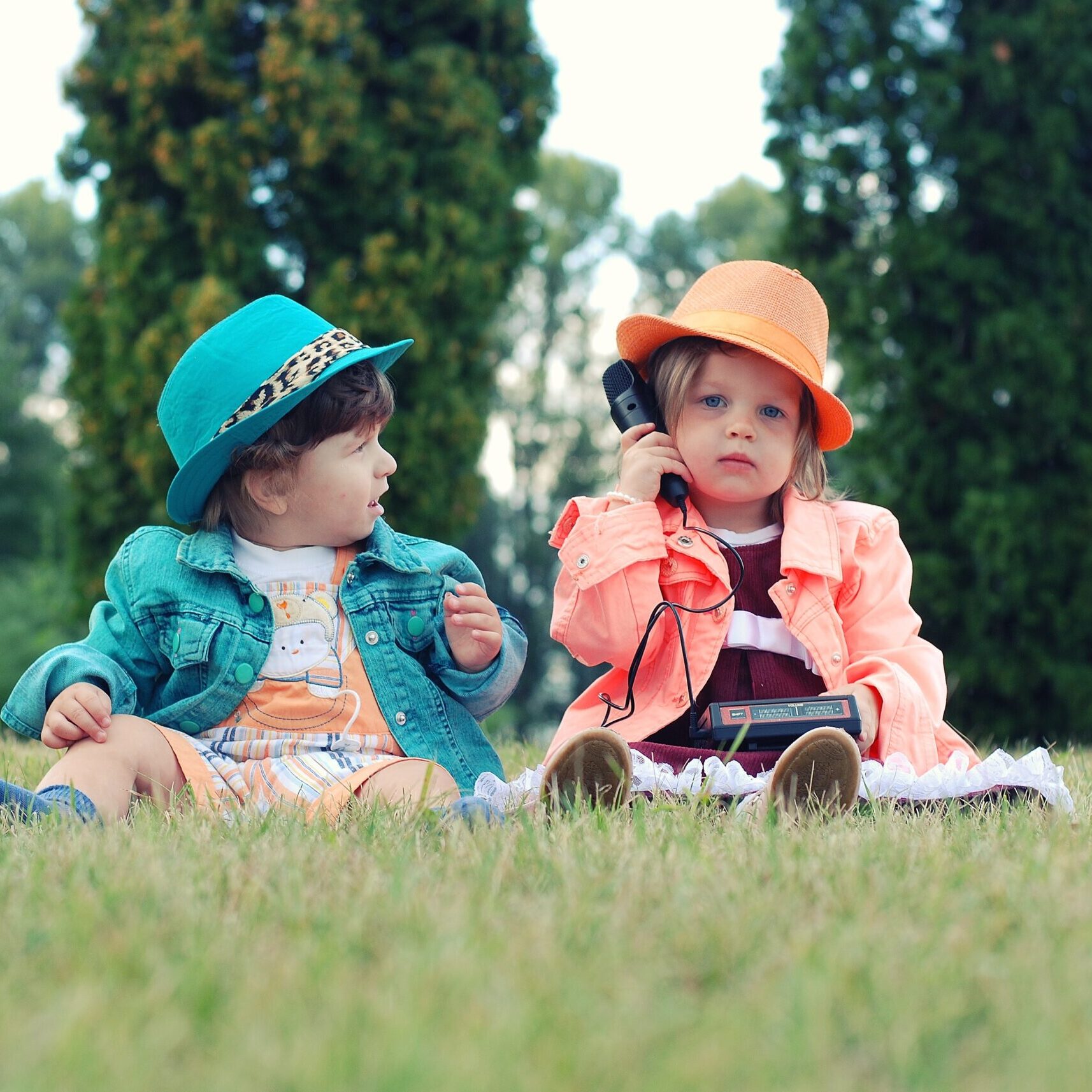 Two very young girls sitting on the grassy knoll as one appears to be holding a microphone and the other girl looks at her in amazement.